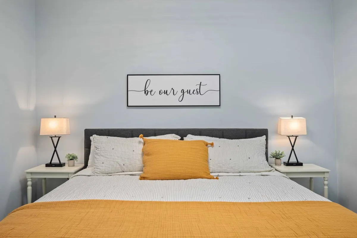 A cozy bedroom setup with a comfortable bed, decorative pillow, wall art, nightstands, and table lamps. The warm color tones create a restful and inviting space for sleeping or relaxing.