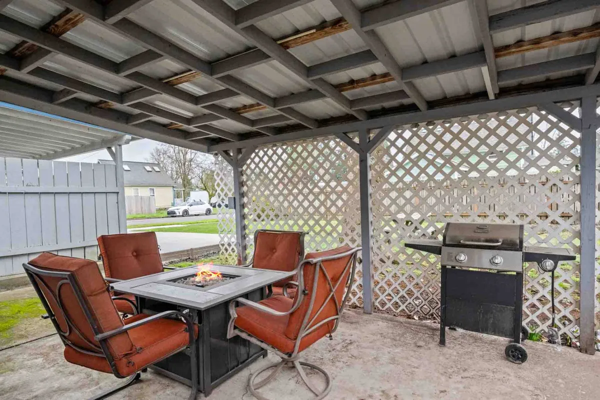 A covered patio at dusk featuring a fire pit table surrounded by comfortable chairs, with a grill in the background and a relaxed atmosphere perfect for social gatherings. The scene captures guests enjoying sunset views while dining and making s'mores by the fire.