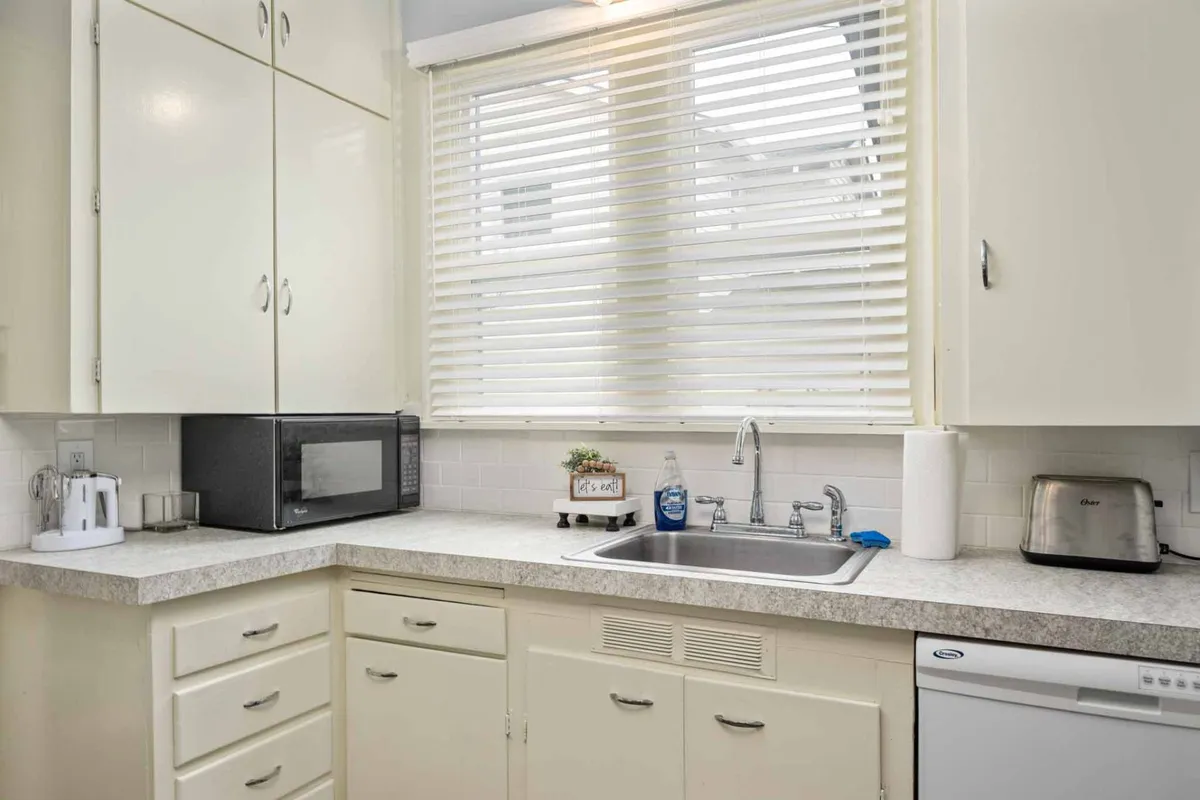 A bright, inviting kitchen with morning light streaming through a window with blinds. The kitchen features a sink, microwave, and cabinets, suggesting a hub for meal prep and casual gatherings. The window provides a glimpse of the outdoors, hinting at the home's connection to the surrounding environment.