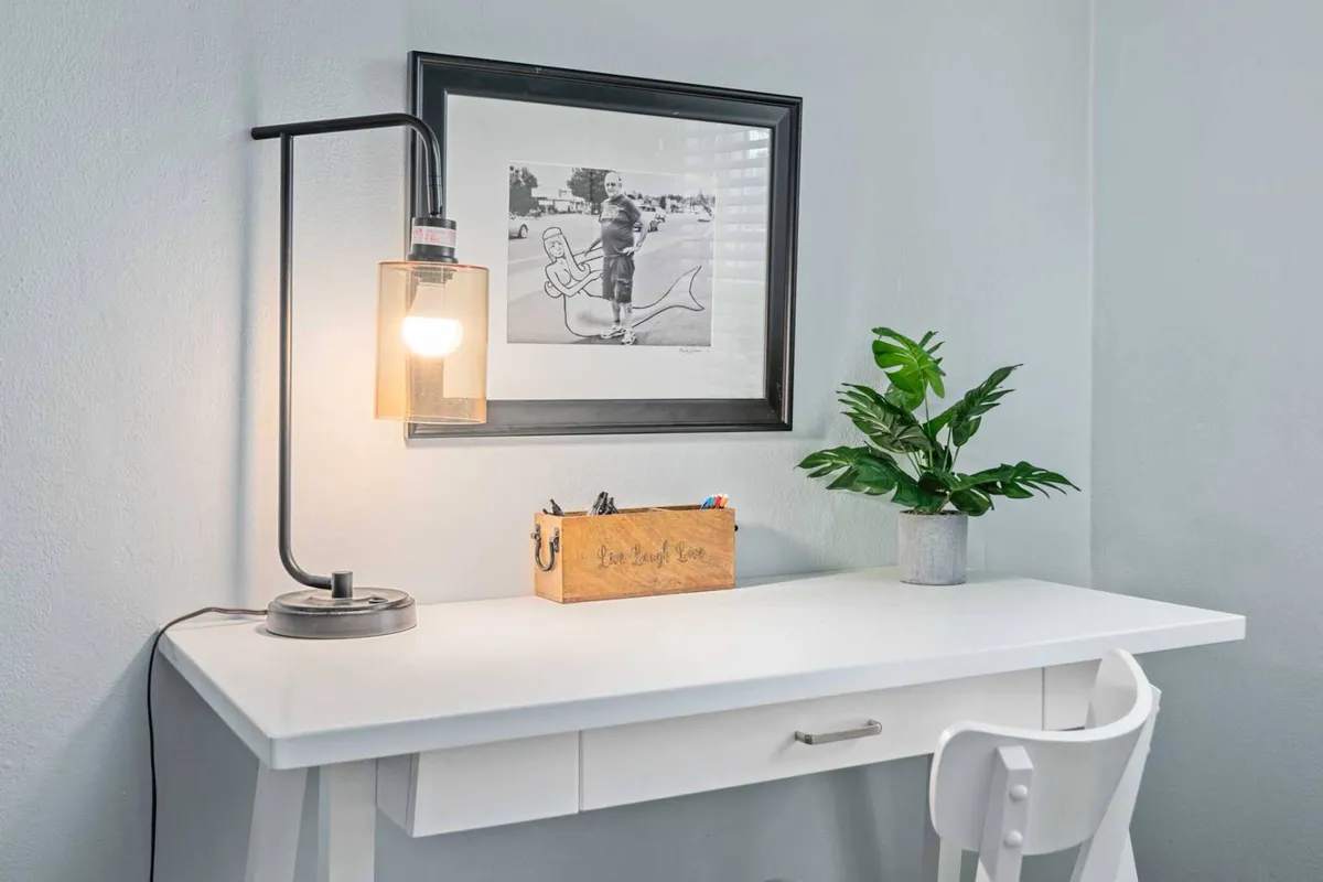 A peaceful workspace illuminated by a desk lamp, featuring a framed photograph, potted plant, and a wooden box. The setting is creative and contemplative, hinting at moments of reflection or creativity that tie into the area’s storied past.