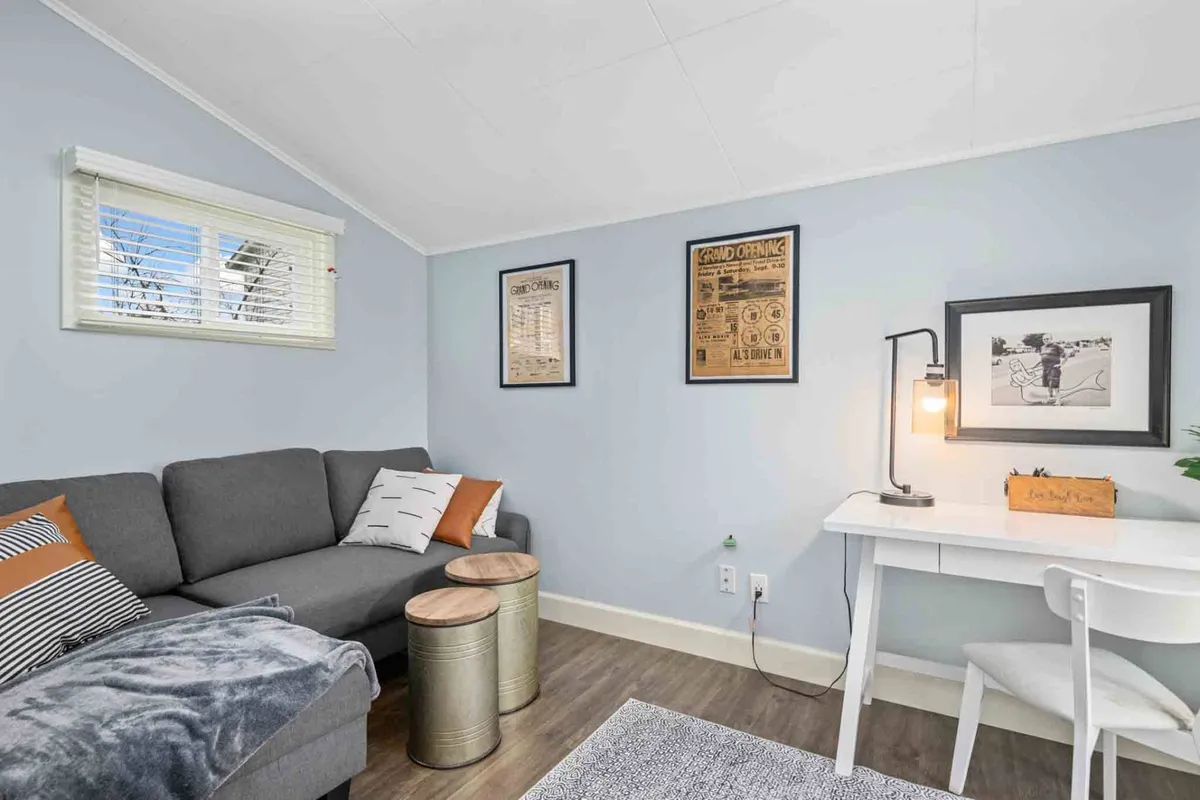 An inviting office space featuring a gray couch with decorative pillows, wooden side tables, wall artwork, and a desk with a lamp. The dual-purpose nature of the room is evident, making it suitable for work, relaxation, or even as a sleeping space with the queen sleeper sofa.