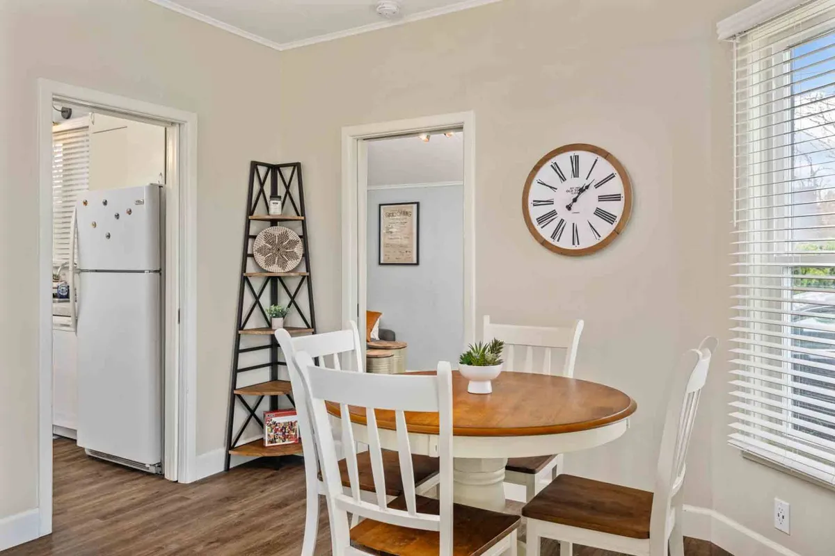 This image displays a round wooden dining table surrounded by white chairs, a wall clock, and a decorative shelf. The cozy and inviting atmosphere is perfect for family meals, casual gatherings, or even working on school assignments together.
